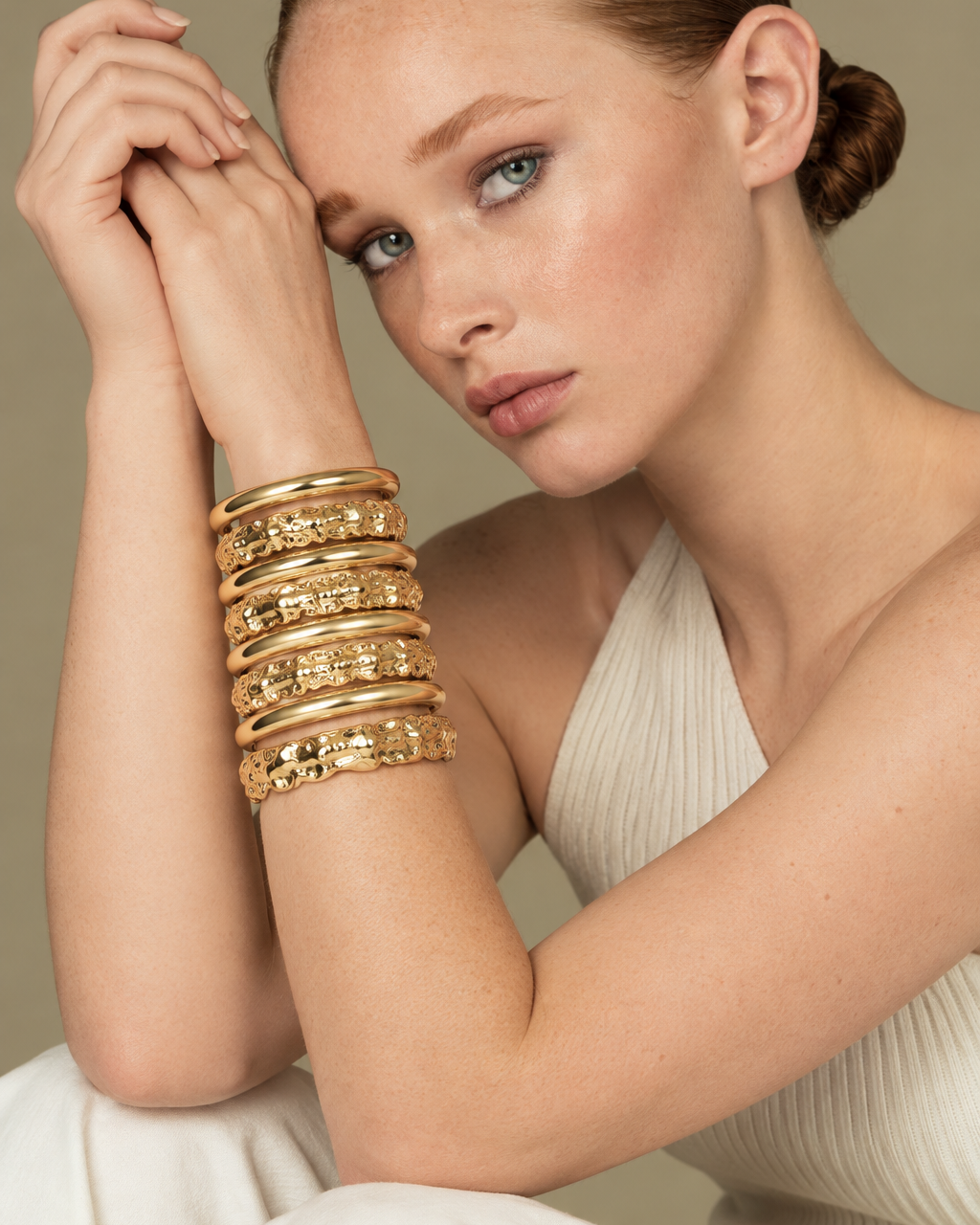 Close-up of a woman resting her head on her hands, wearing the Nathalia gold cuff bracelet stacked with other gold bangles, cream sweater
