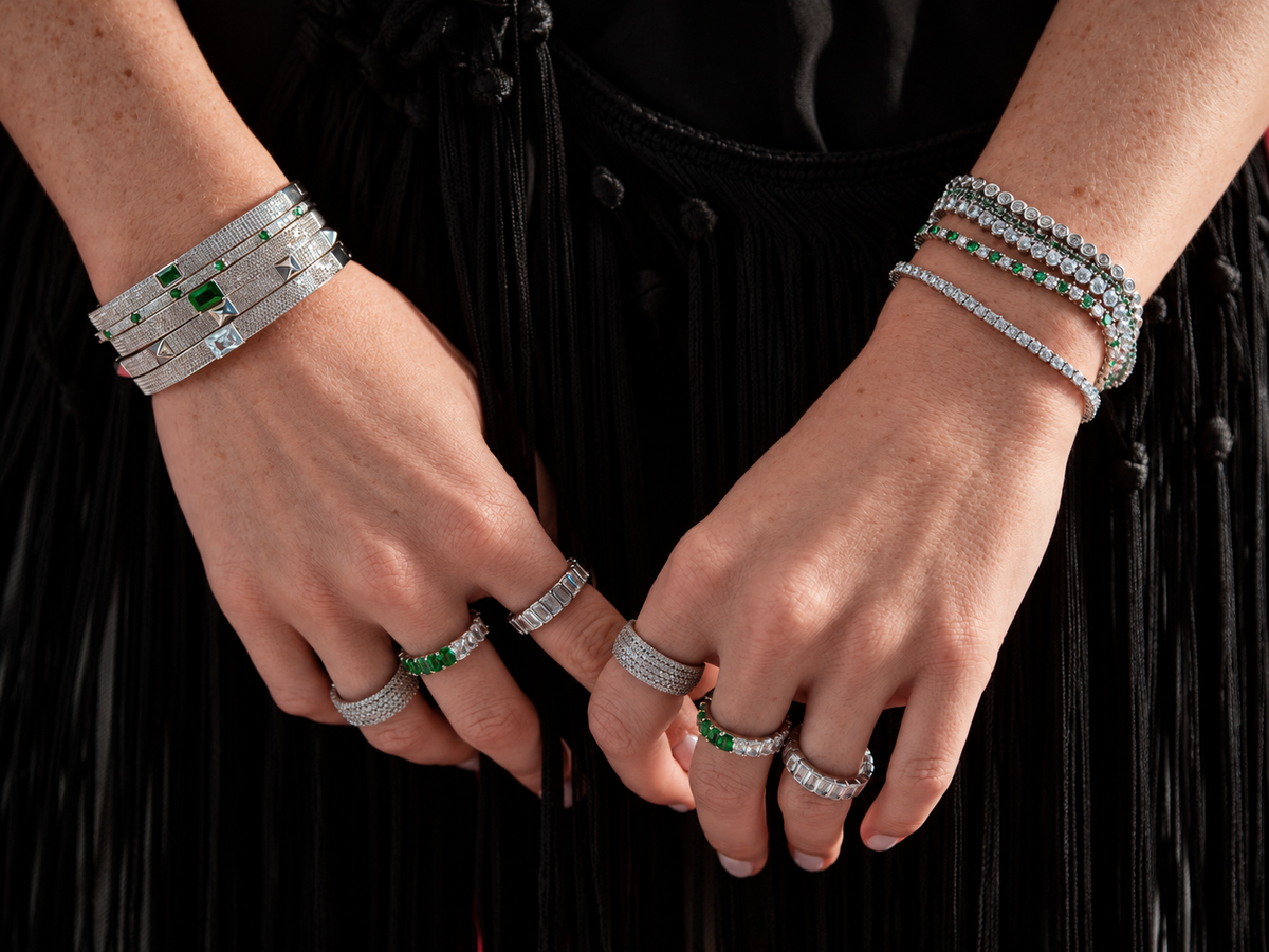 Lifestyle photo of two crossed wrists in dark clothing, styled with multiple bangle and tennis bracelets stacked together along with rings.