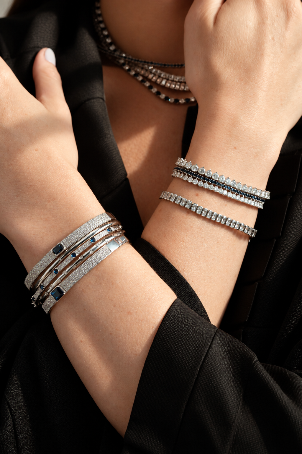 Lifestyle photo of two crossed wrists in black clothing, styled with multiple bangle and tennis bracelets stacked together, some layered with a beaded necklace.