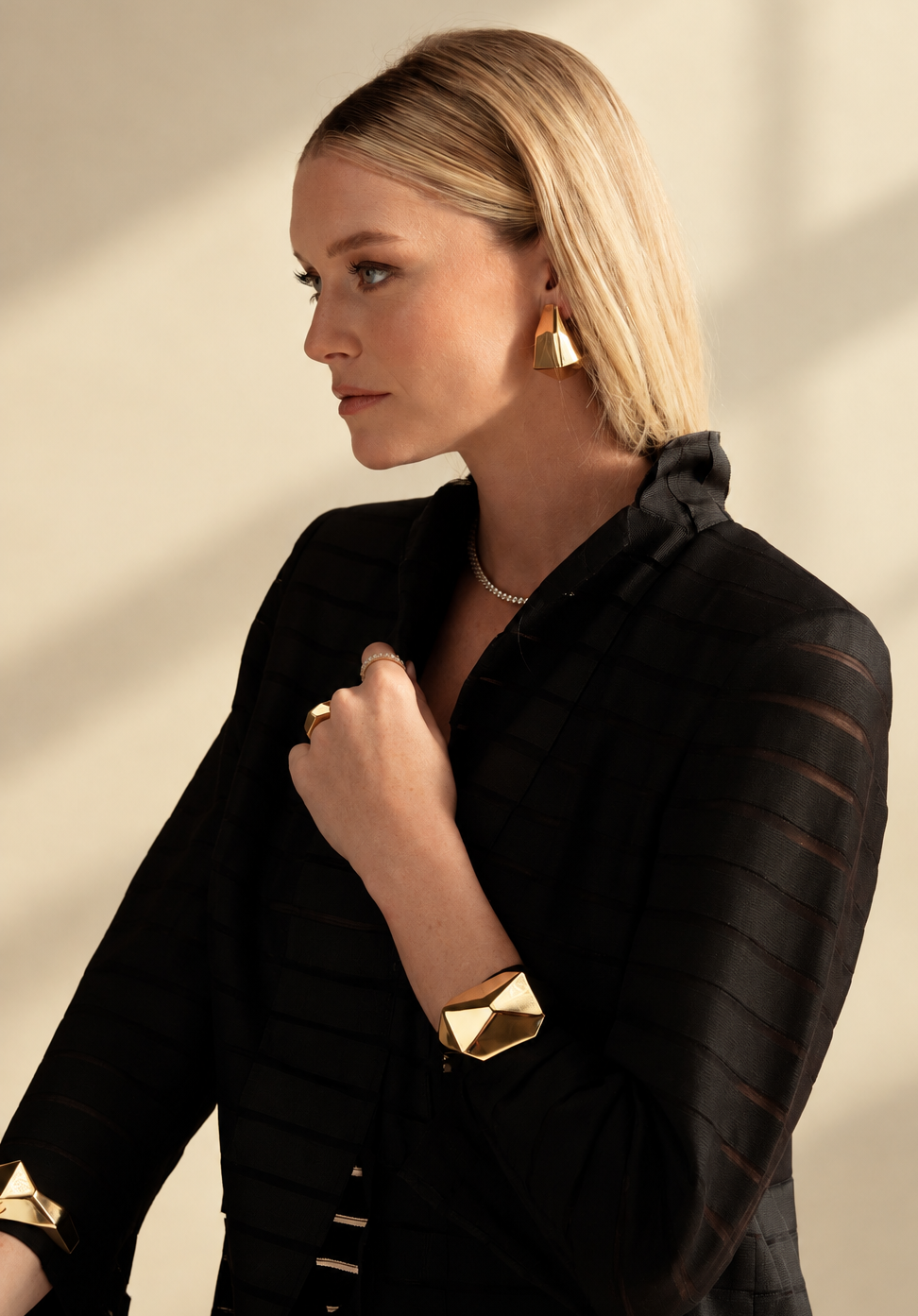 Lifestyle photo of a woman in a black top with her hand near her chest, wearing the gold geometric cuff bracelet and earrings.
