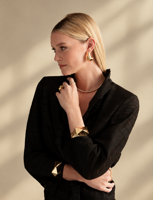 Lifestyle photo of a woman in a black top with her hand near her chin, styled with gold geometric chunky hoop earrings and a gold geometric cuff bracelet worn together.