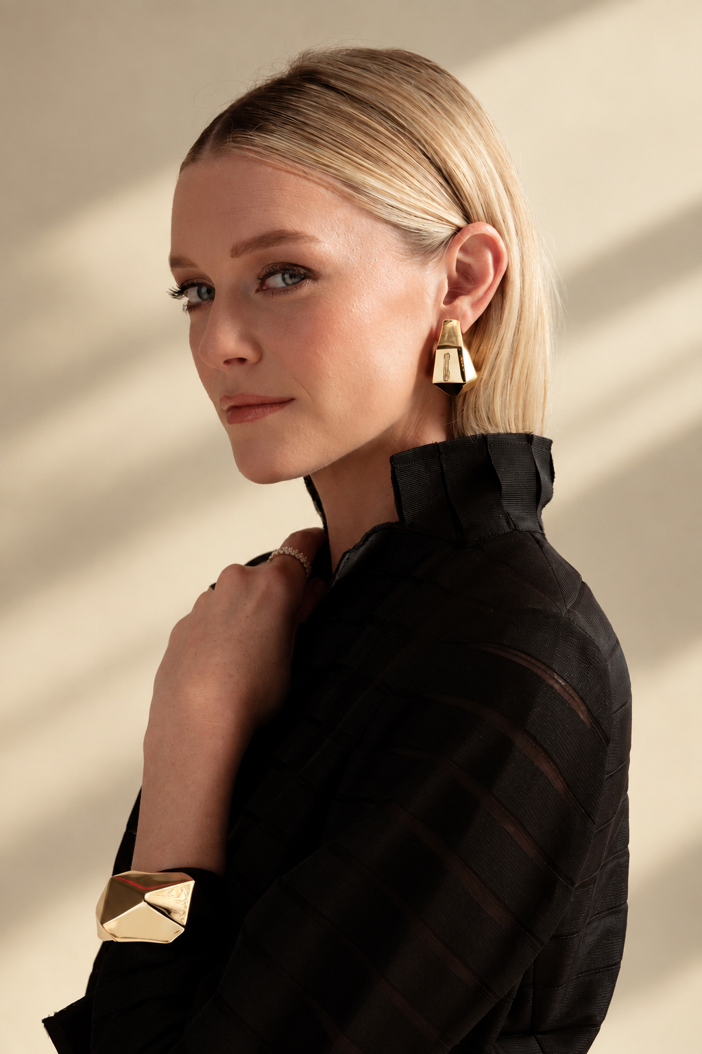 Lifestyle photo of a woman in a black top looking over her shoulder, styled with gold geometric chunky hoop earrings and a gold geometric cuff bracelet worn together.