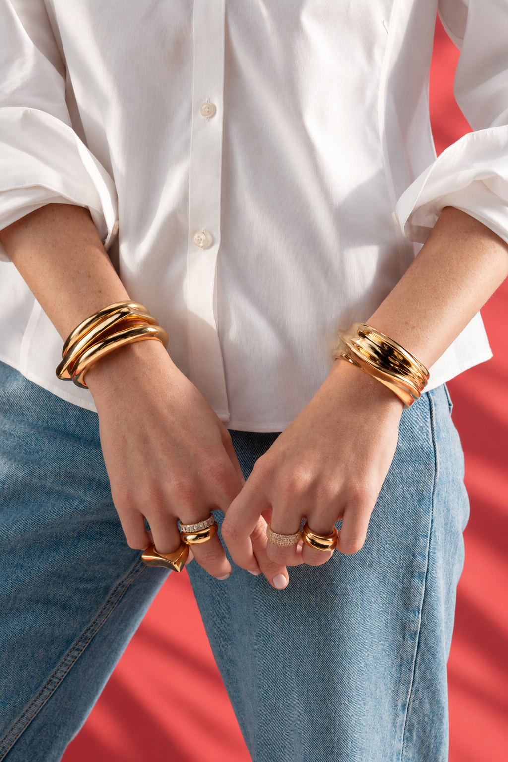 Lifestyle photo of a woman in a white shirt and jeans against a red background with her hands near her waist, wearing the chunky sculptural ring and stacked bangles.