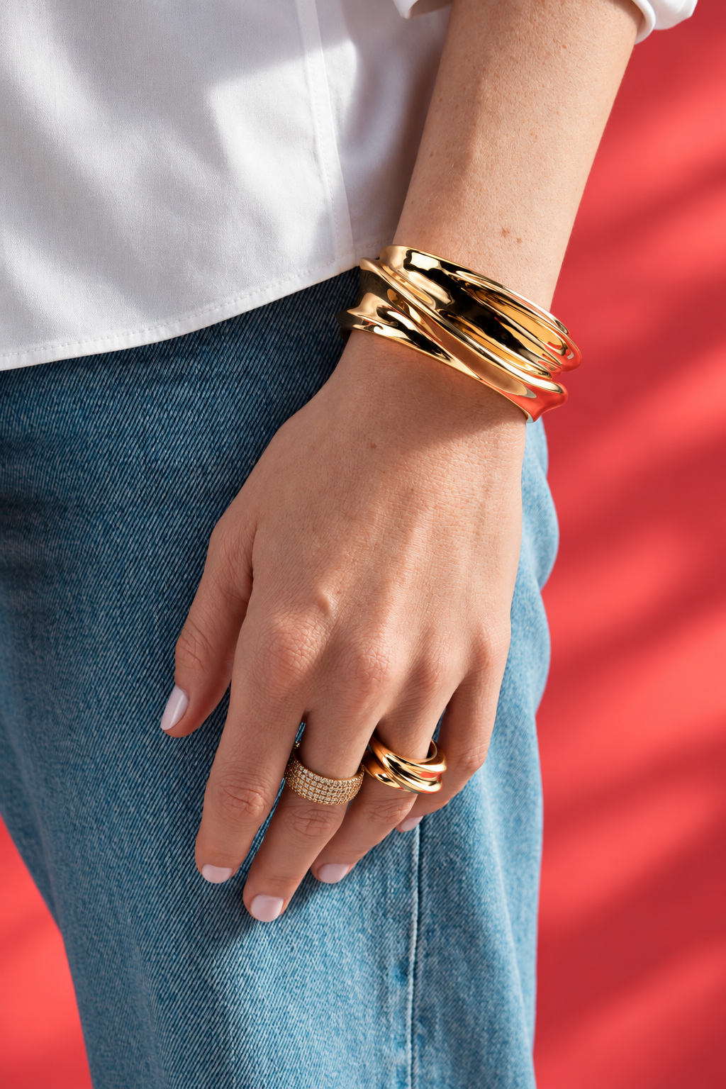 Lifestyle close-up of a hand resting on denim jeans against a red background, wearing the chunky sculptural ring and a bangle.