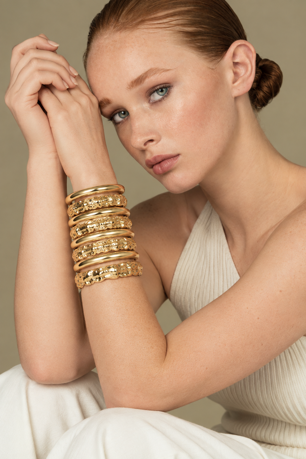 Close-up of a woman resting her head on her hands, wearing the Nathalia gold cuff bracelet stacked with other gold bangles, cream sweater