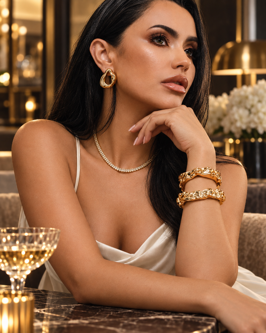 Close-up of a woman resting her cheek on her hand, wearing the Nathalia gold cuff bracelet stacked with other bracelets, a necklace and earrings, in a restaurant setting with a glass of wine