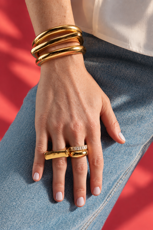 Close-up of a wrist stacked with gold bangles and the Caroline gold statement ring, red background