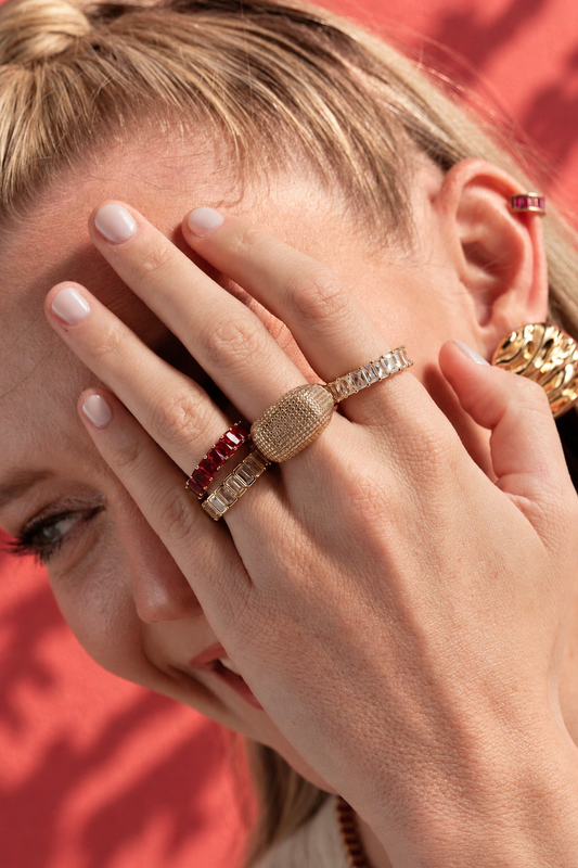Lifestyle photo of a woman against a red background with her hand covering part of her face, styled with a wide band pave ring stacked with other rings, along with a multicolor baguette ear cuff.
