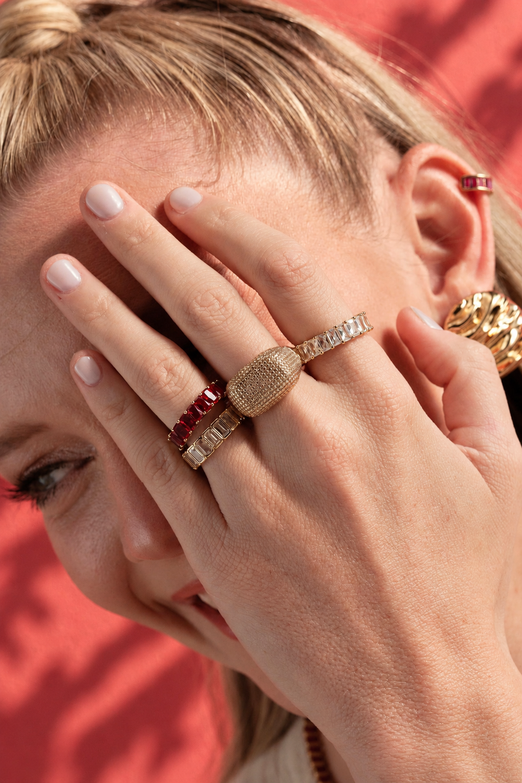 Lifestyle photo of a woman against a red background with her hand covering part of her face, styled with multiple rings stacked together, including an eternity ring.