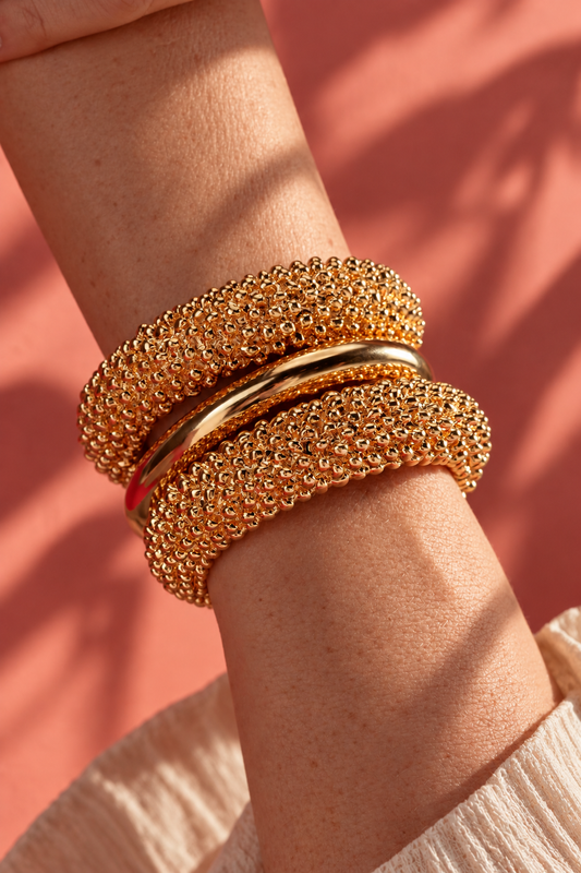 Close-up of a wrist stacked with textured gold Isabella bangles against a warm orange background
