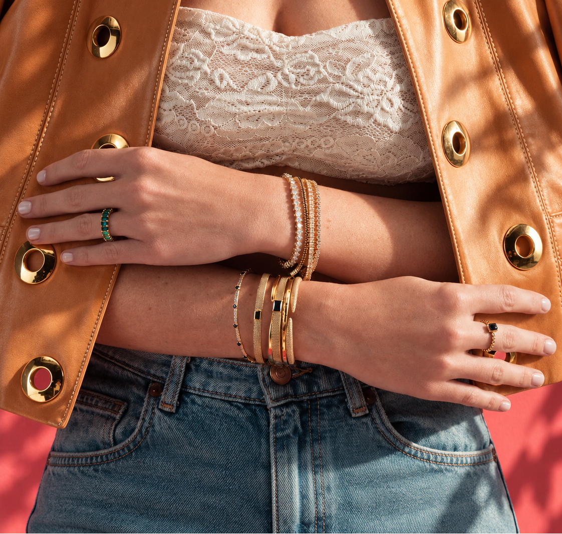 Lifestyle close-up of a hand against caramel leather, styled with multiple bangle and cuff bracelets stacked together with rings.