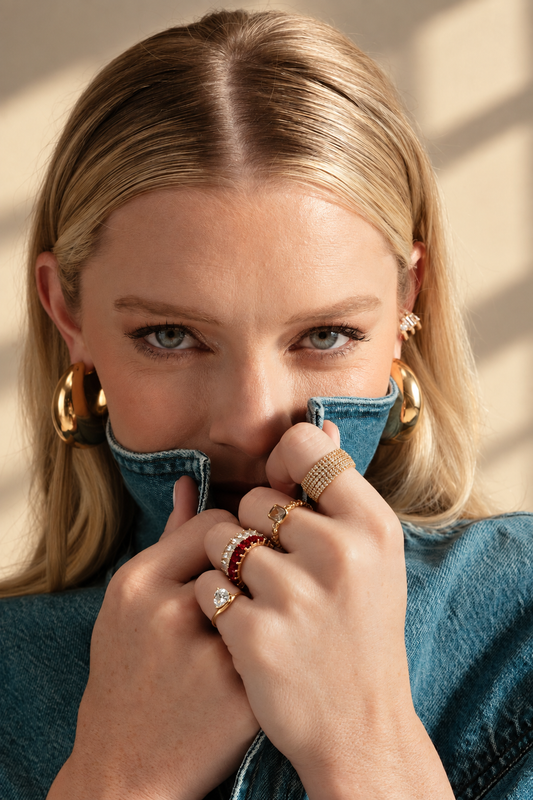 Lifestyle photo of a woman in a teal top against a red background with her hands near her face, styled with multiple rings worn together, including an eternity ring, a band ring, and a pinky ring.
