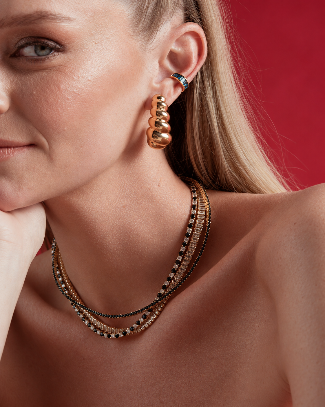 Close-up of a woman's neck and ear, wearing the Catarina gold hoop earrings layered with gold necklaces, red background