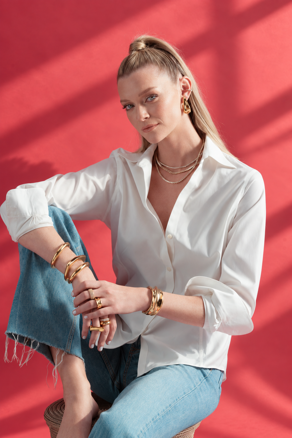 Woman sitting cross-legged wearing a white shirt and jeans, with the Sophia silver cuff bracelet stacked with gold bangles, red background