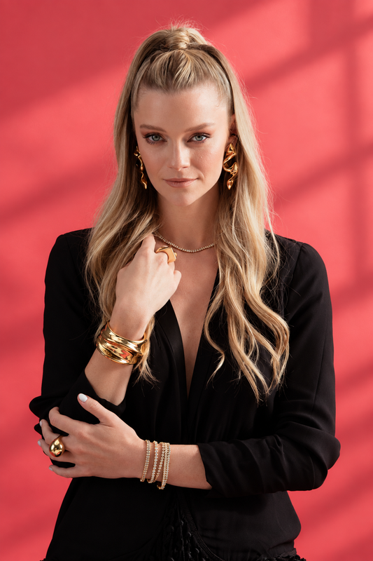 Lifestyle photo of a woman in a black top against a red background with her hand near her collarbone or chest, styled with a gold cuff bracelet, a gold spiral statement drop earring, rings, and a necklace layered together.