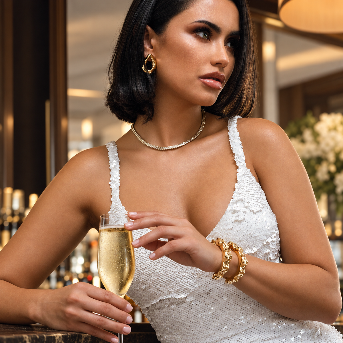 Woman in a white dress holding a glass of champagne, wearing the Nathalia gold cuff bracelet stacked with other bracelets, restaurant setting