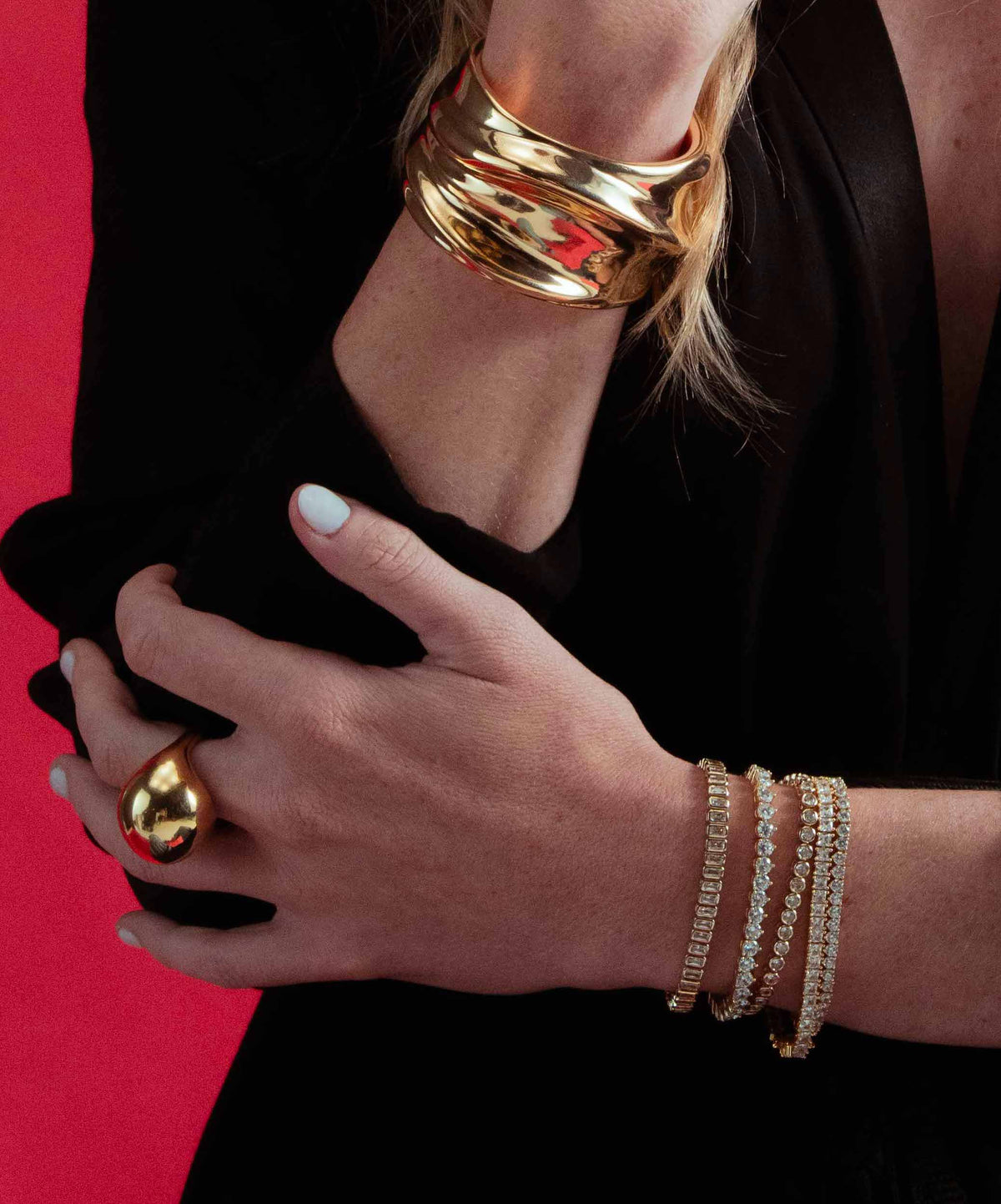 Close-up of a wrist stacked with the Laura gold tennis bracelet and gold bangles, hand resting against the wrist, red background