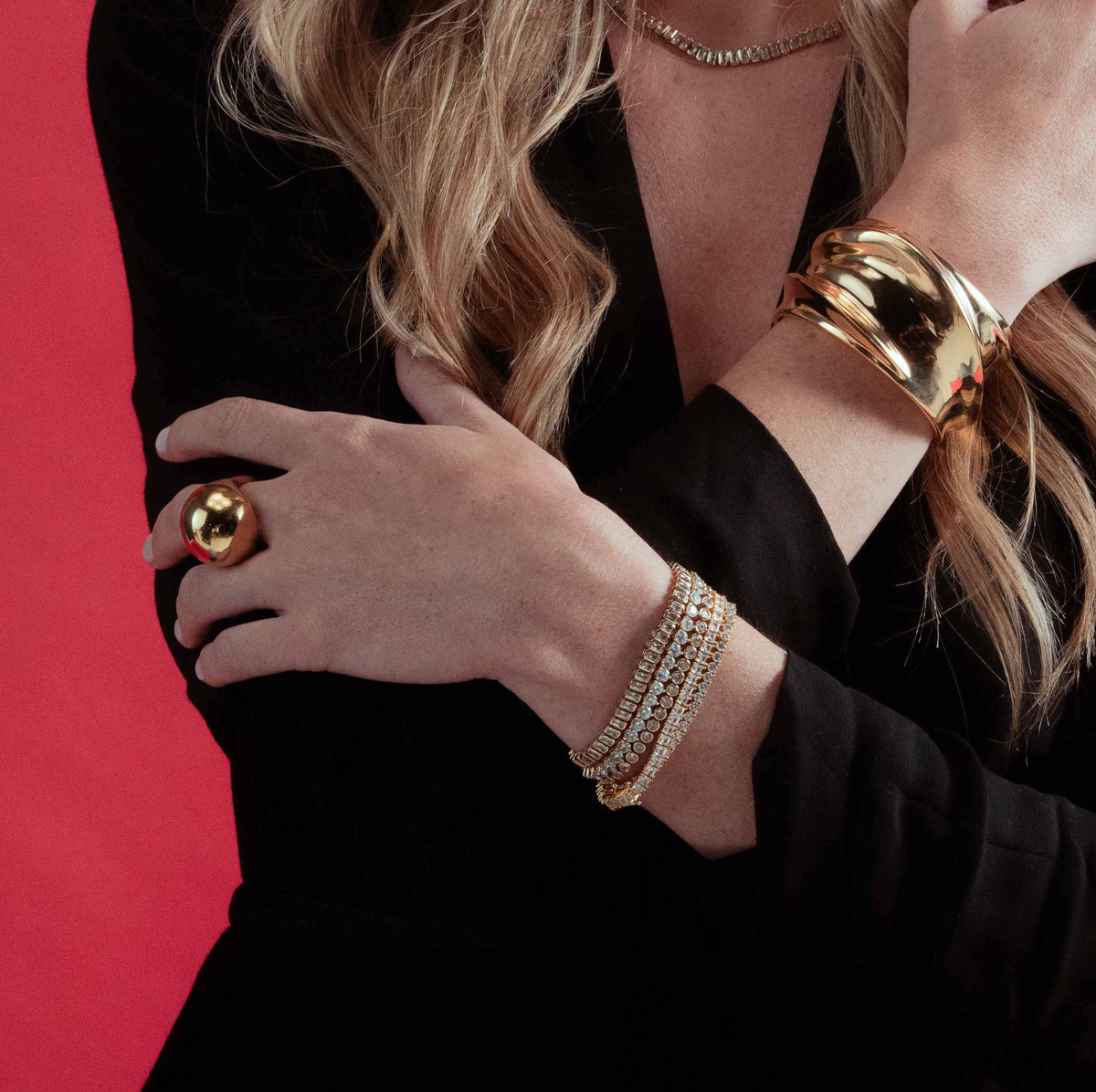 Lifestyle photo of a woman with her hand near her chin, wearing the gold wide cuff bracelet stacked with a ring and other bracelets, against a red background.
