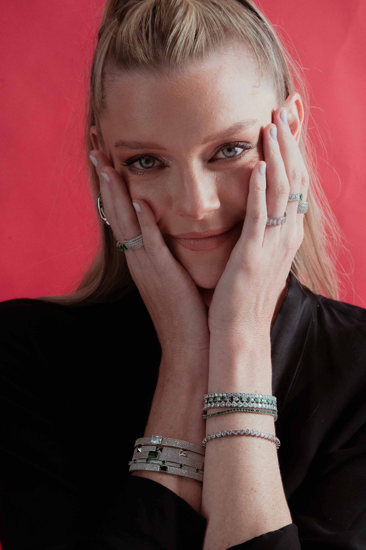 Lifestyle photo of a woman in a black top with her hands near her face, styled with a tennis bracelet layered together with other bracelets and rings, against a dark/red background.