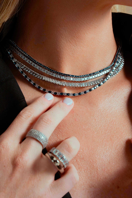 Close-up of a woman's chest wearing layered necklaces including the Laura silver tennis choker, with a ring on her hand near her chin, black top