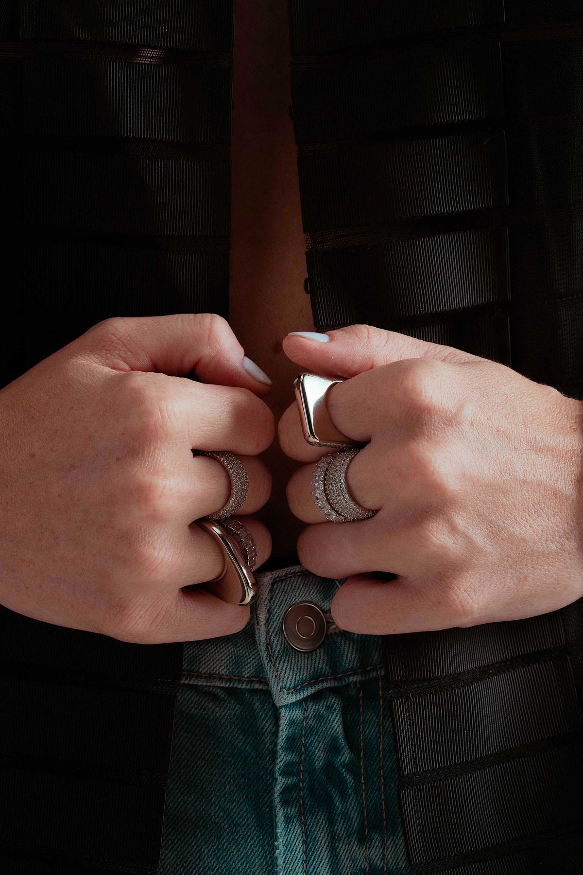Close-up of two hands resting near a waistband, wearing the Lily gold signet ring stacked with other rings, dark denim