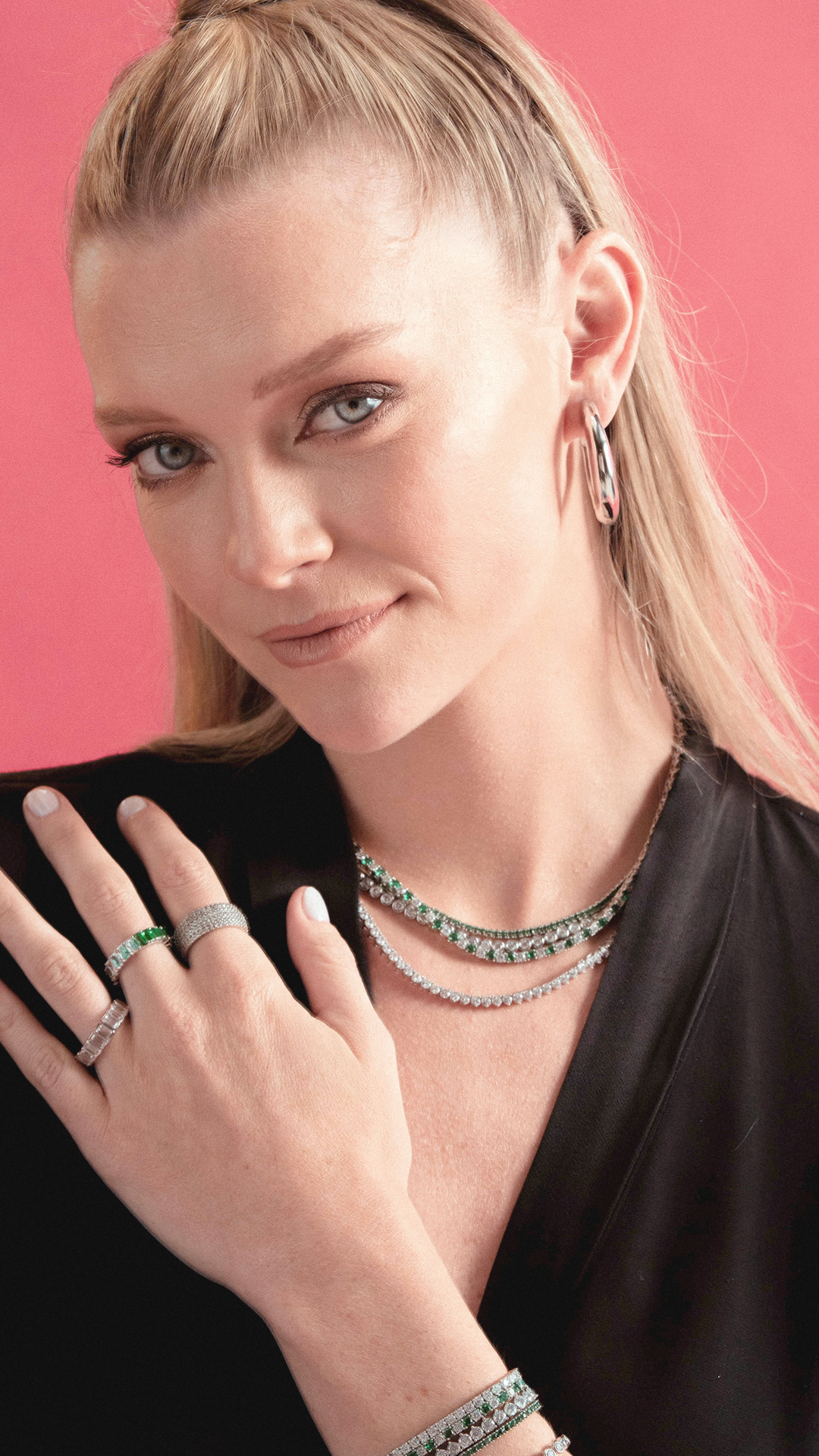 Woman wearing gold-tone earrings, layered necklaces, and multiple rings featuring simulated emeralds and simulated diamonds, against a coral pink background.