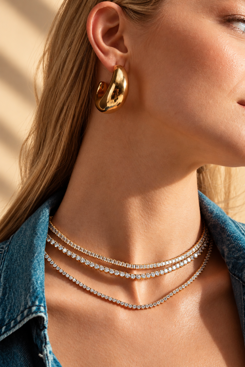 Lifestyle close-up of a woman's ear in a denim jacket, wearing a sculptural earring along with the three-prong tennis necklace layered with other necklaces.