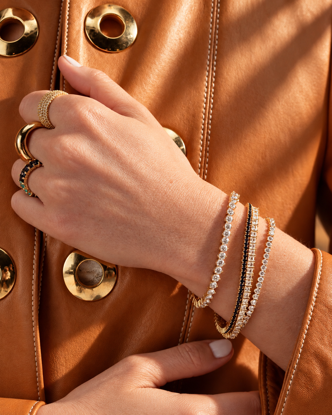 Lifestyle close-up of a hand against caramel leather, styled with a tennis bracelet stacked together with other bracelets and rings.