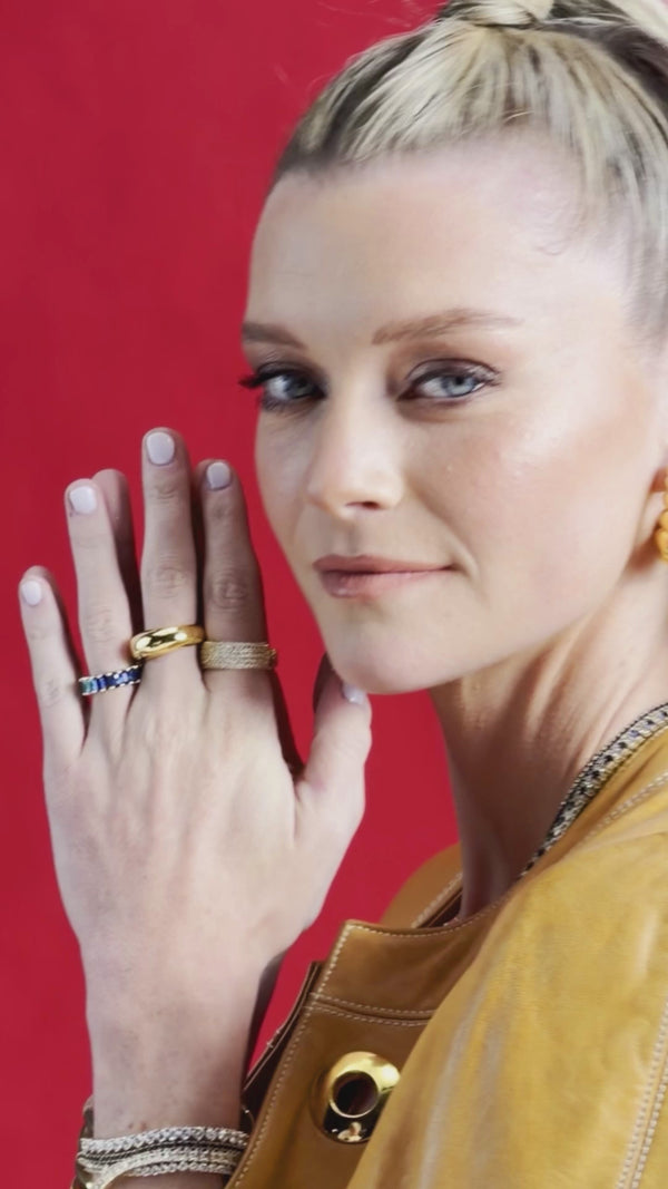 Woman showcasing gold-tone rings, including a blue stone ring, with layered necklaces and gold-tone earrings against a red background.