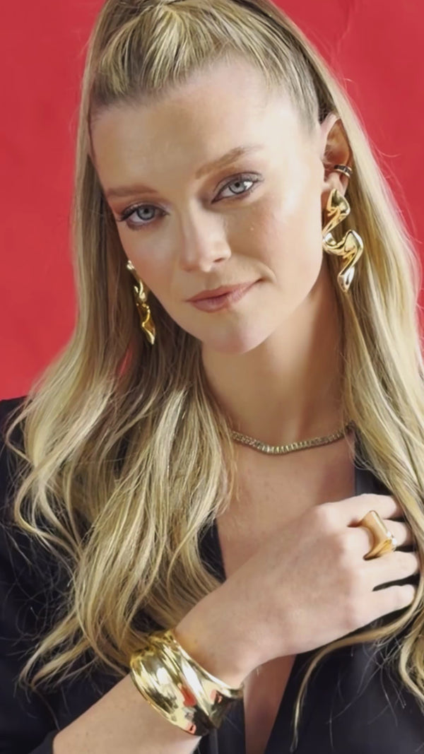 Woman wearing sculptural gold-tone drop earrings, a delicate gold-tone necklace, and a ring against a red background.