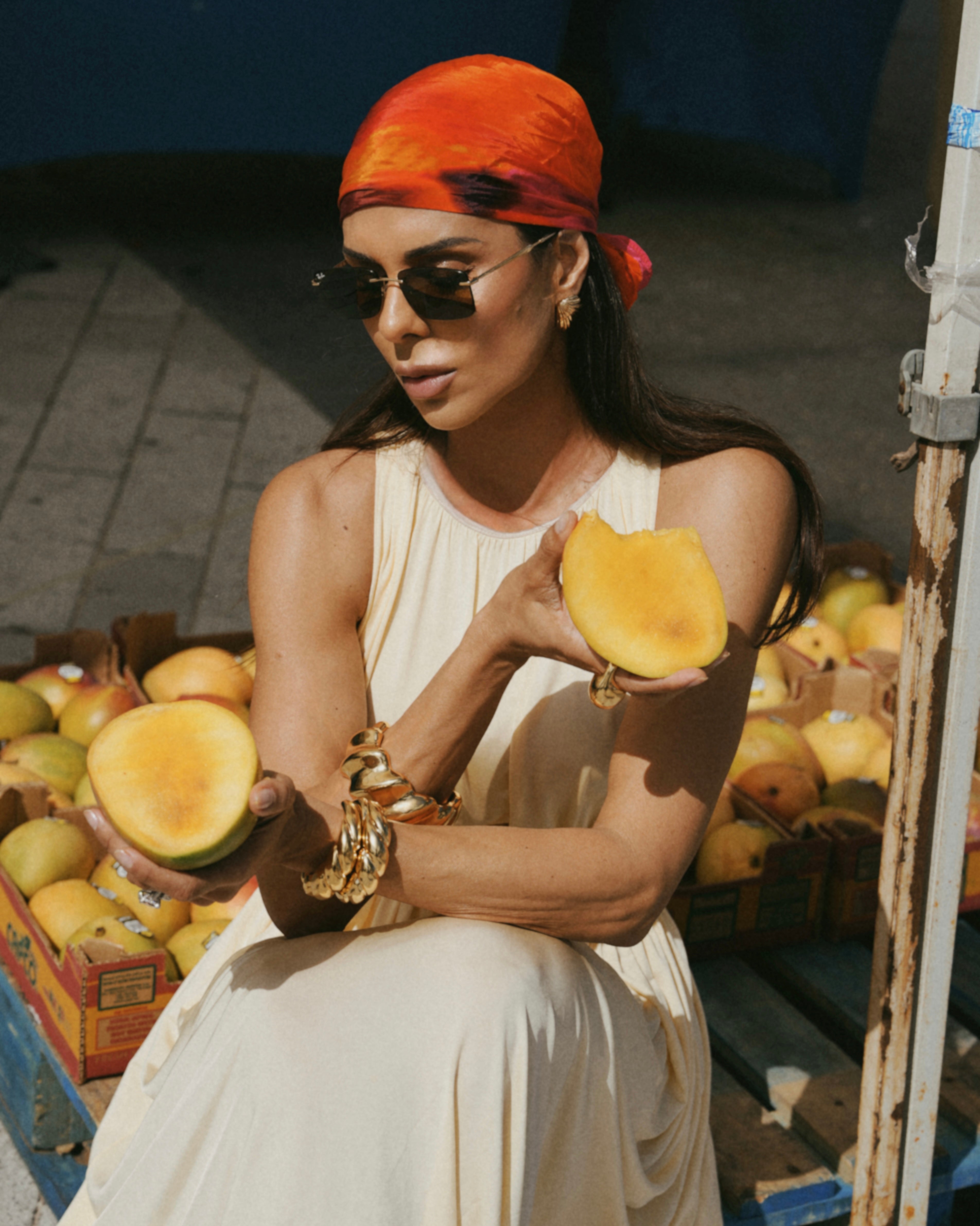 A woman wearing a cream sleeveless dress, gold bracelets, and a bright orange headscarf sits outdoors holding two halves of a fresh yellow mango. Boxes of mangoes are visible in the background.
