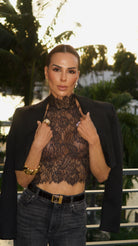 A woman posing outdoors wearing a sheer black lace mock-neck crop top under an open black blazer, paired with dark gray jeans and gold jewelry.